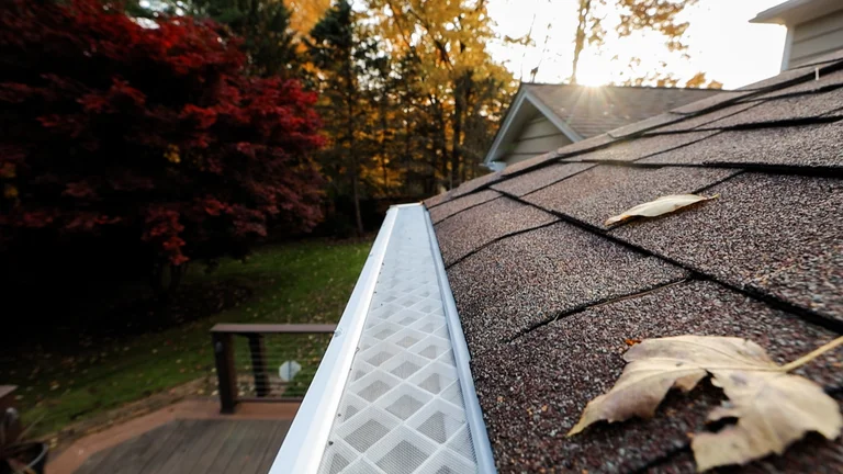 White micromesh gutter guard running along a shingle roof edge with autumn leaves resting on top and Upstate fall foliage behind